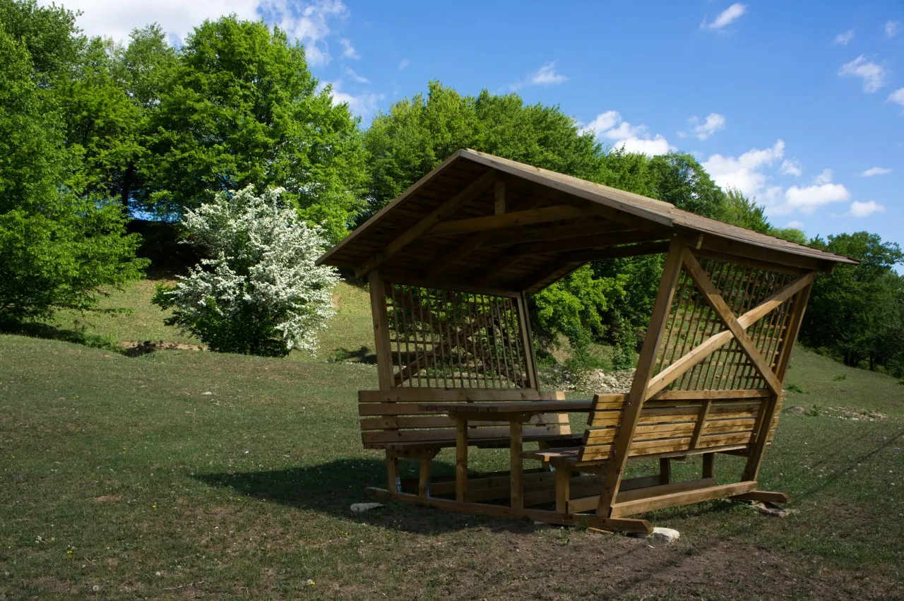 A timber gazebo with a shingle roof and lattice sides, with benches and a table, in a mown clearing; behind it a white flowering tree and beech woods.
