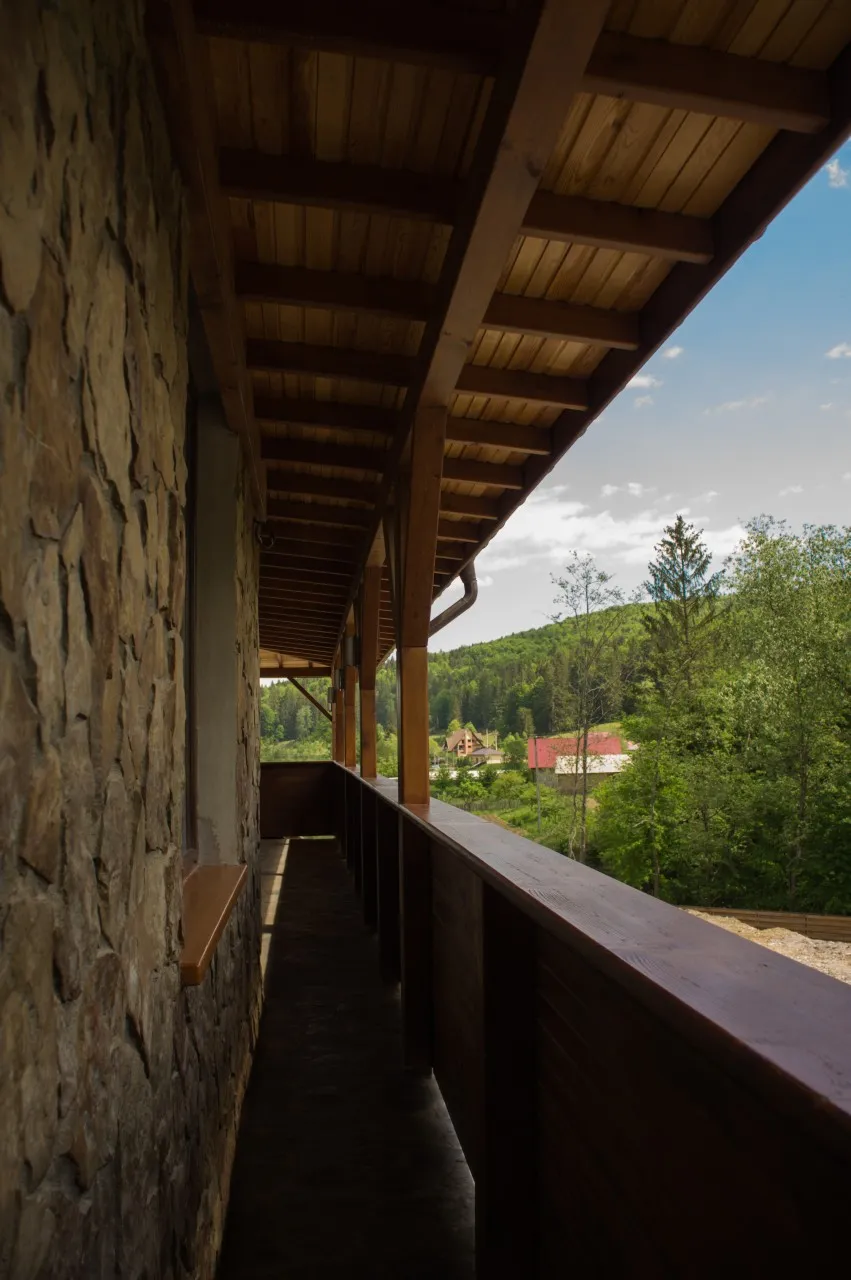 A covered balcony running along the building, with a stone wall and wooden posts, open towards the hills and the village below.