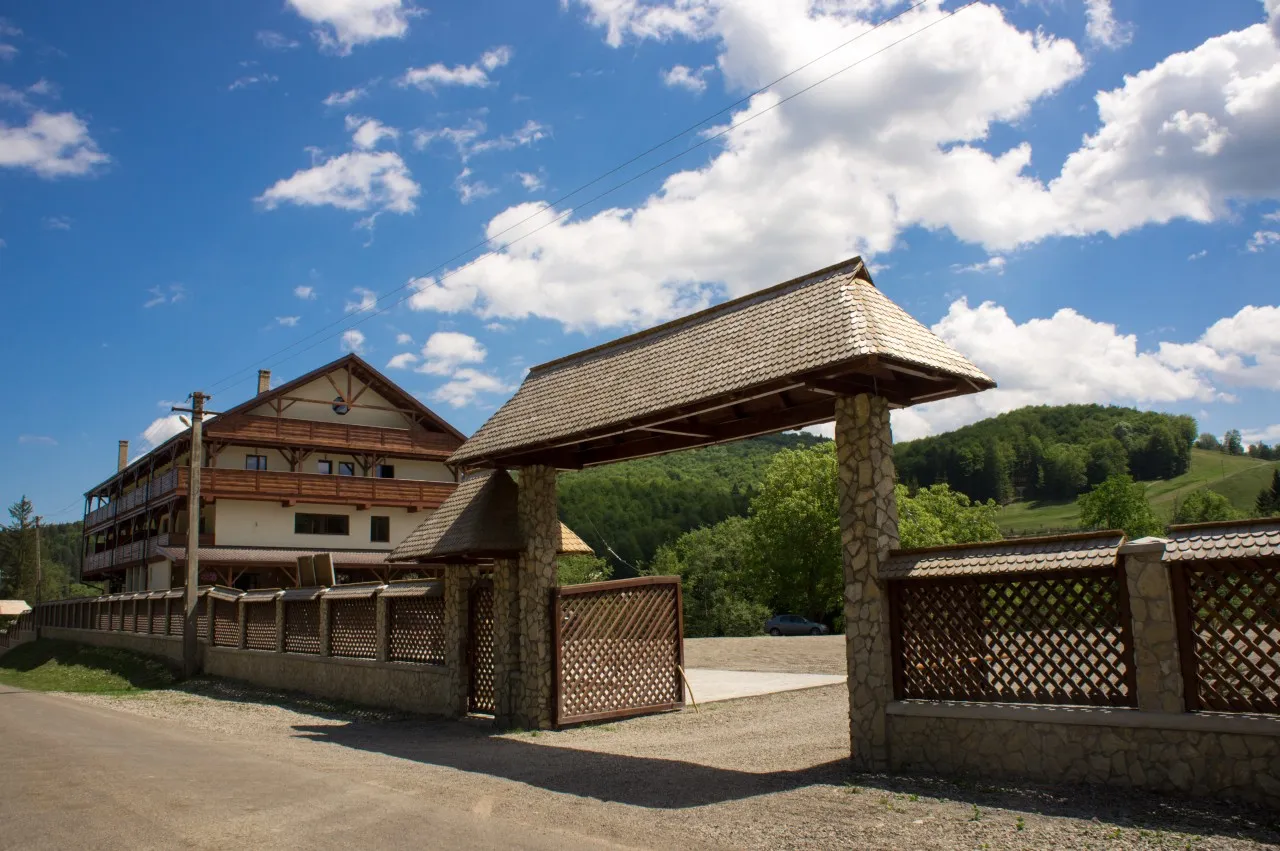The chalet’s gate seen from the road, with stone piers, a timber leaf and a shingle roof, the building to the left and the wooded hill behind.