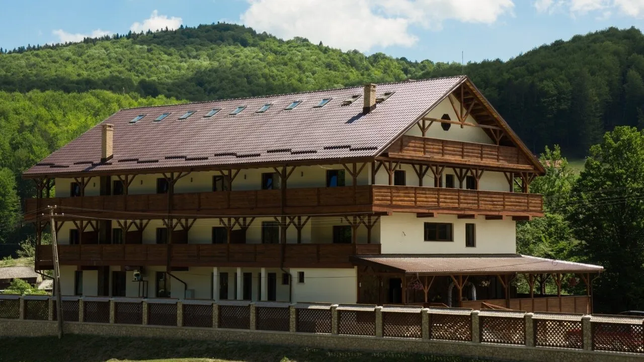 The chalet seen at an angle, with timber balconies across three storeys along the front, at the foot of a wooded hillside.