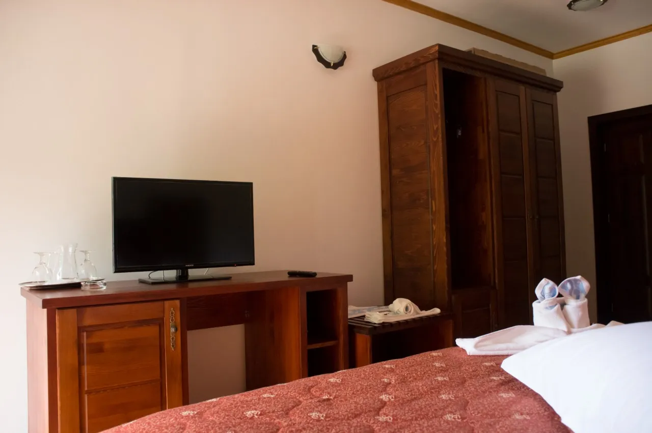 A corner of the room with a TV on a wooden dresser, a tall wardrobe and the edge of the bed