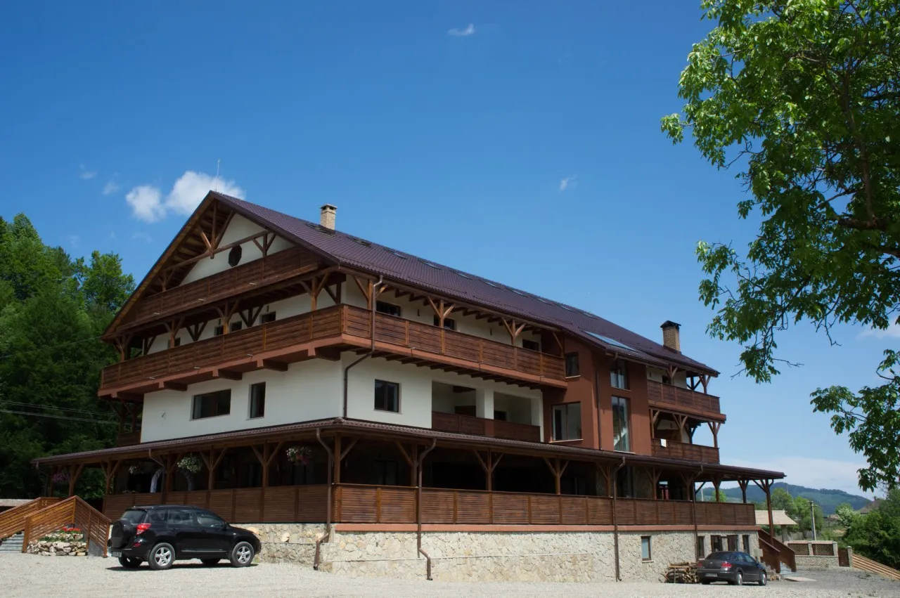 The chalet seen from its yard, with timber balconies along the front, the covered terrace at ground level and the gravel parking in front.