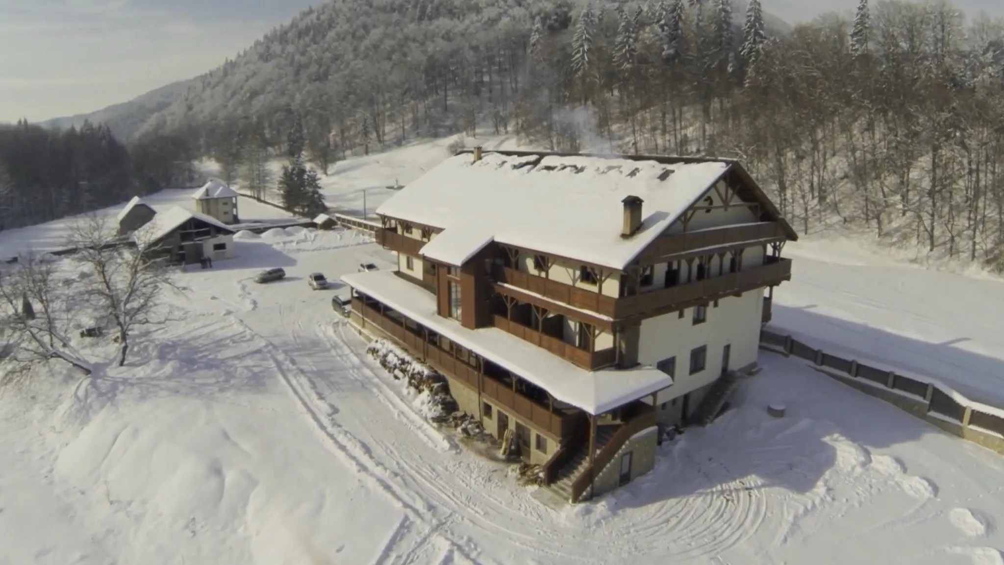 The chalet from the air in winter, from the yard side: the building at an angle with snow on the roof, boot and tyre tracks across the deep snow of the yard, the outbuildings to the left and the forest behind.