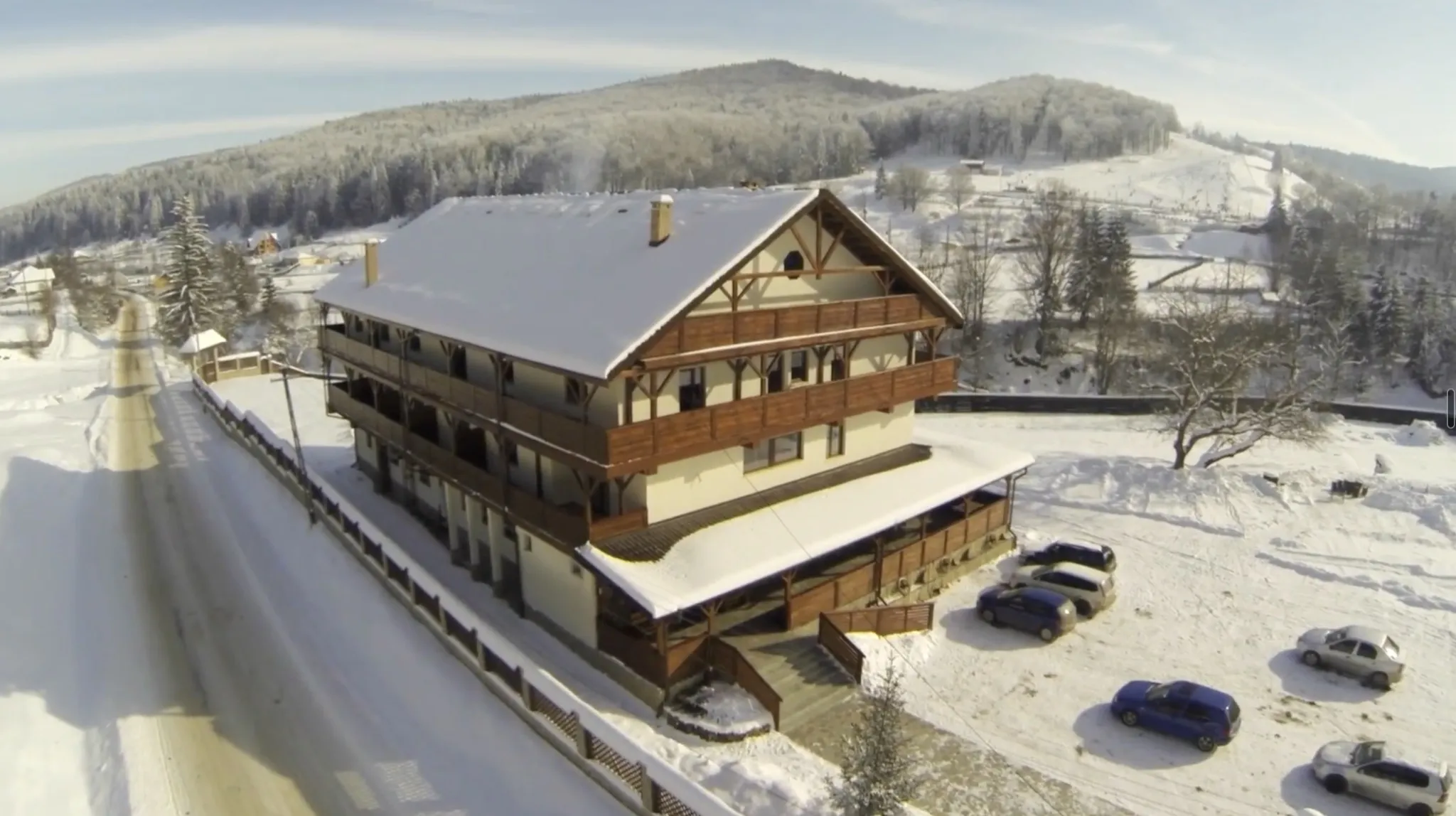 The chalet from the air in winter, from the road side: snow on the roof and on all three tiers of timber balconies, the ploughed road and its fence along the front, snow-covered wooded hills behind.