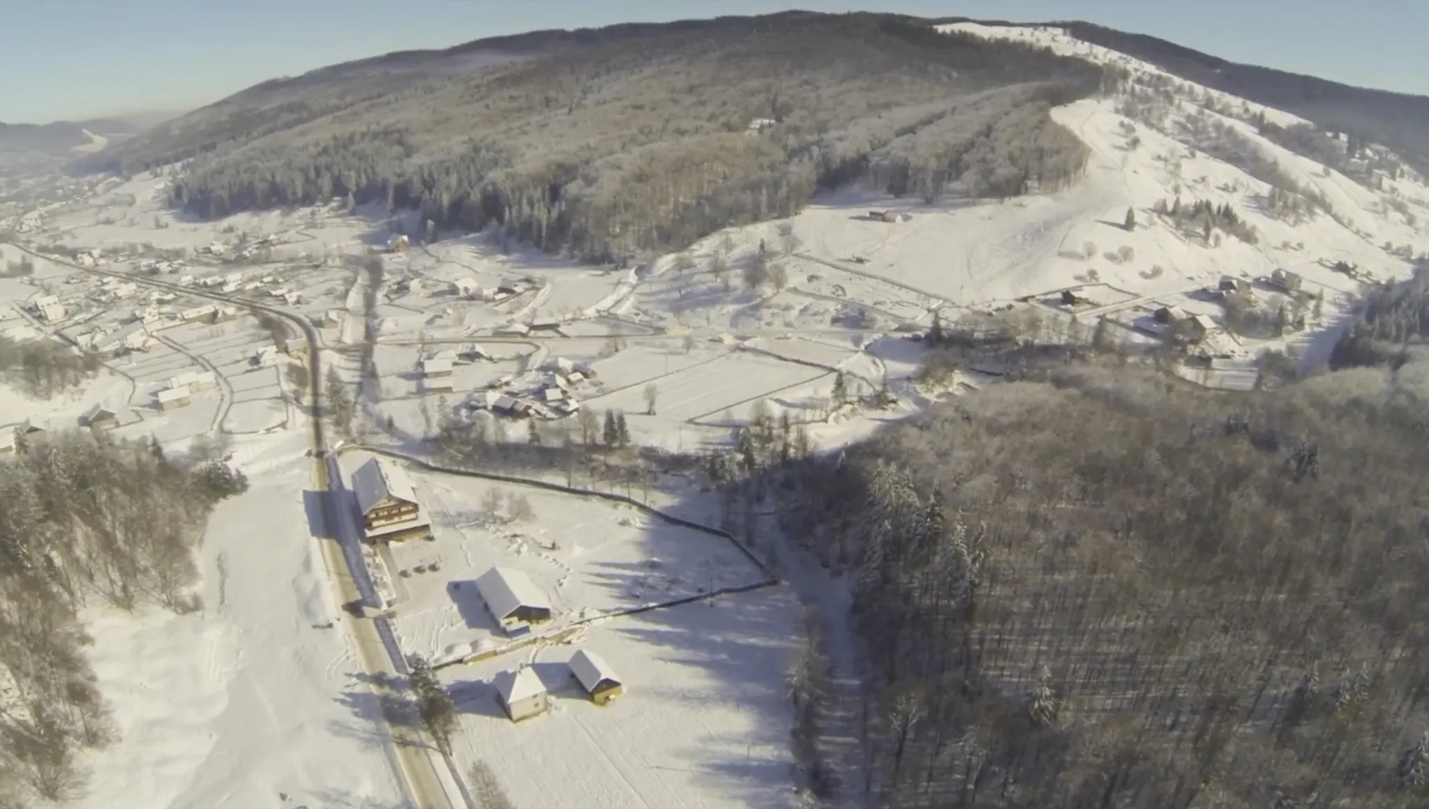 A winter aerial: the chalet and its outbuildings beside the road at the lower left, the snow-covered village in the middle, and the ski slope — a cleared white flank — on the hill to the right.