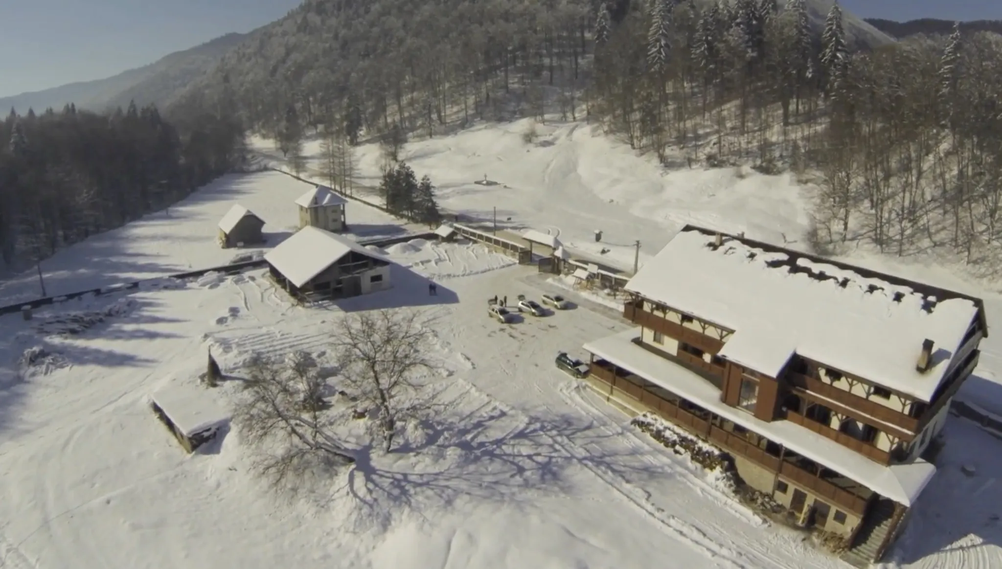 A winter aerial: the chalet at the right, the timber barn and a smaller outbuilding at the left, the swept yard between them and the wooded hillside behind.