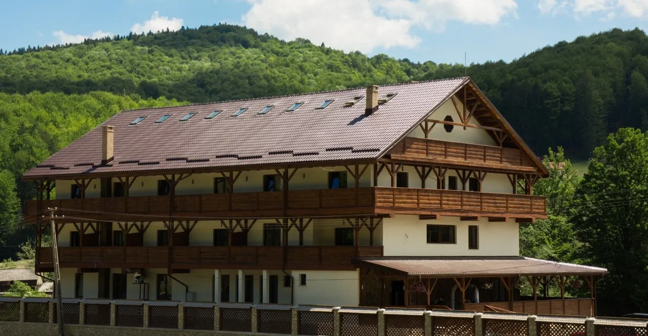 The chalet seen at an angle, with timber balconies across three storeys along the front and the gabled roof end, the wooded hill behind it.