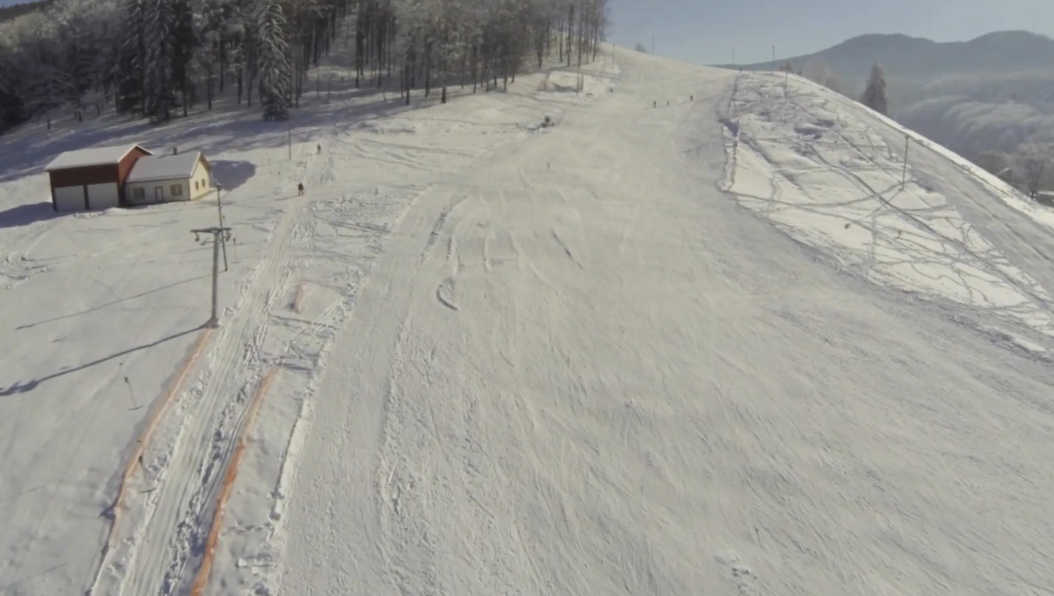 An aerial of the Pojorâta piste: the run rising to the treeline, a ski-tow pylon with its cable at the left, the tow track beside it and a few skiers on the slope. The chalet is not in this frame.