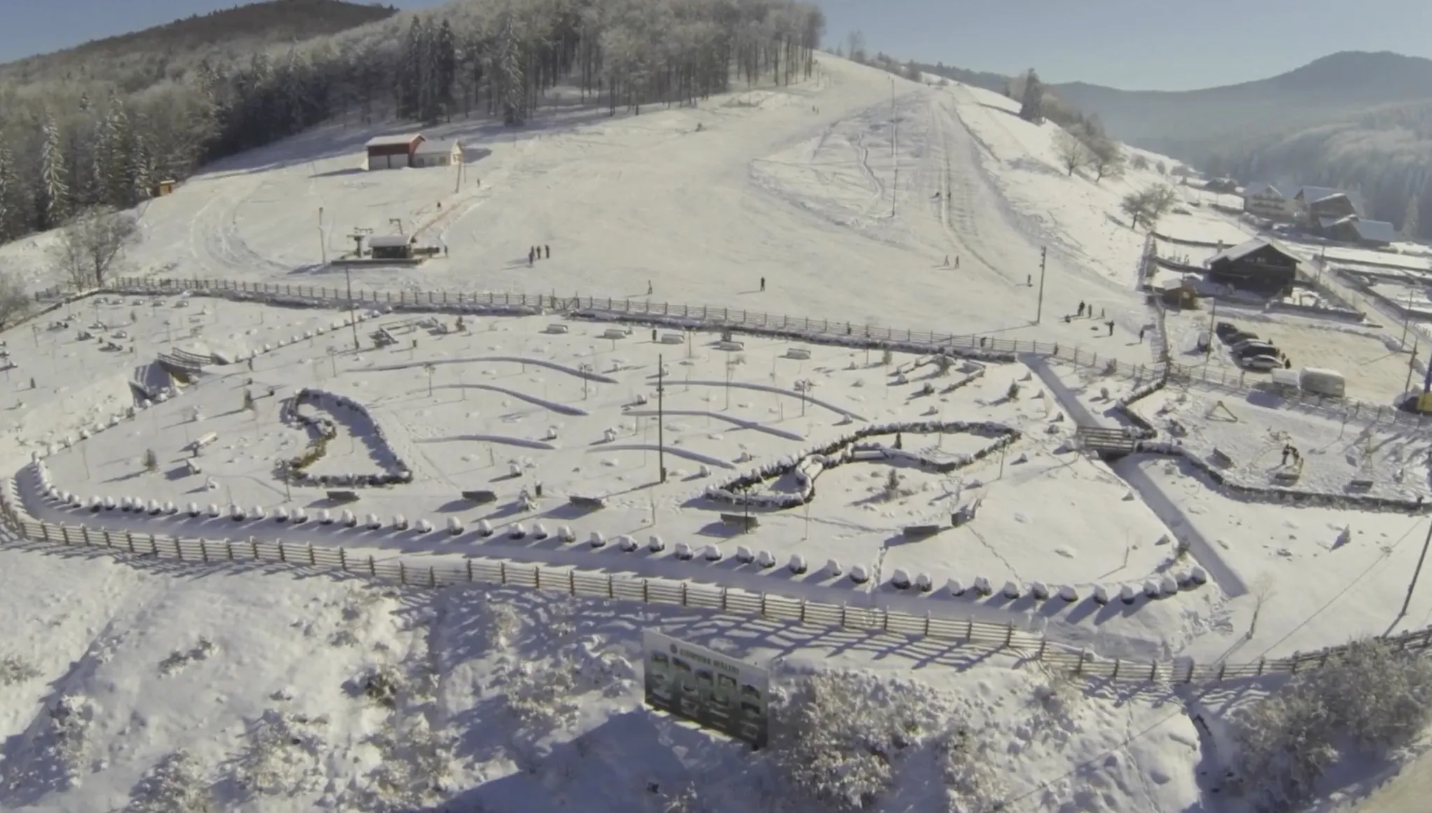 An aerial of the foot of the Pojorâta piste: the ski-tow drive station and the line of pylons climbing the run, a red-roofed hut beside it, people setting off, and in the foreground the fenced park of Mălini commune. The chalet is not in this frame.