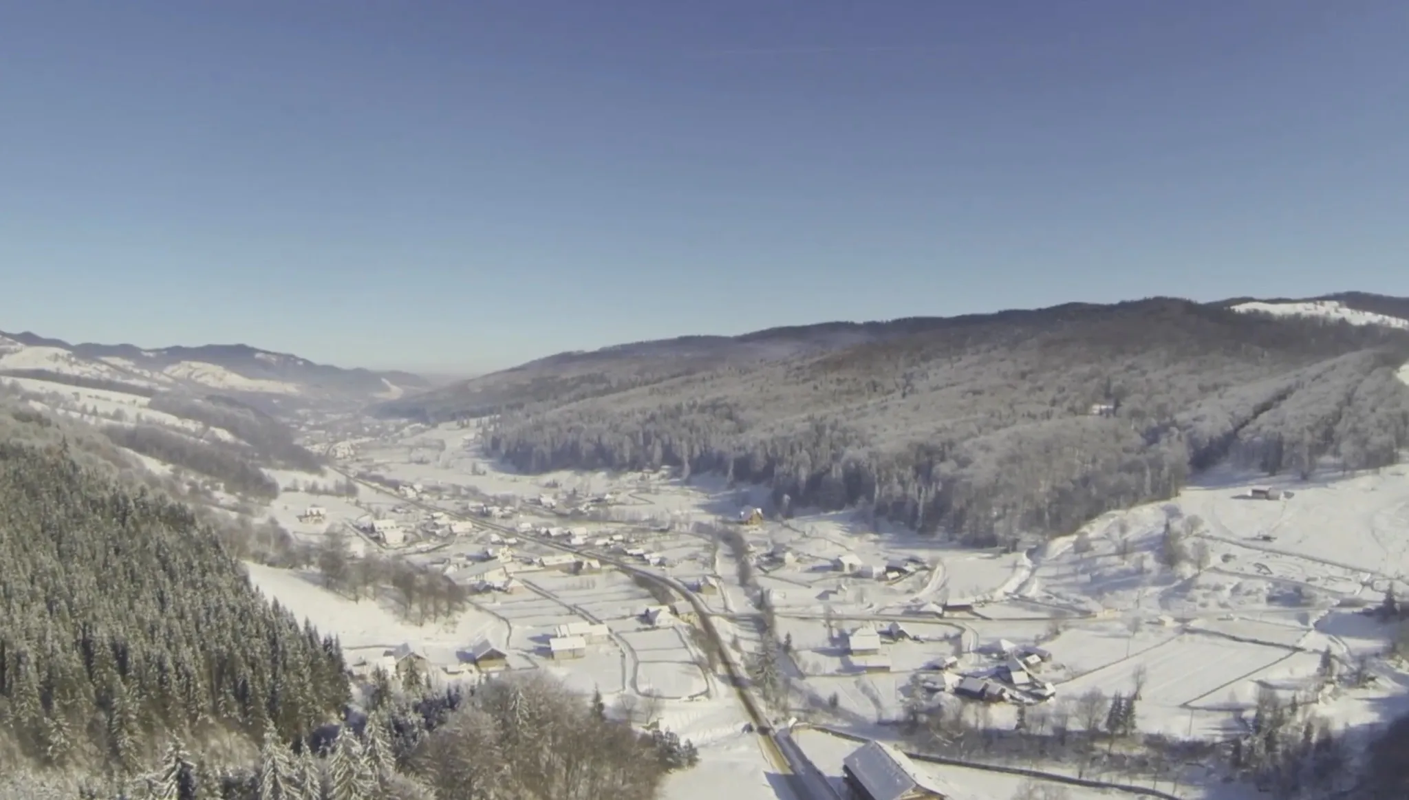 Winter aerial photograph along the length of the valley: the village strung out along the road, strips of snow-covered field, and wooded ridges on either side.